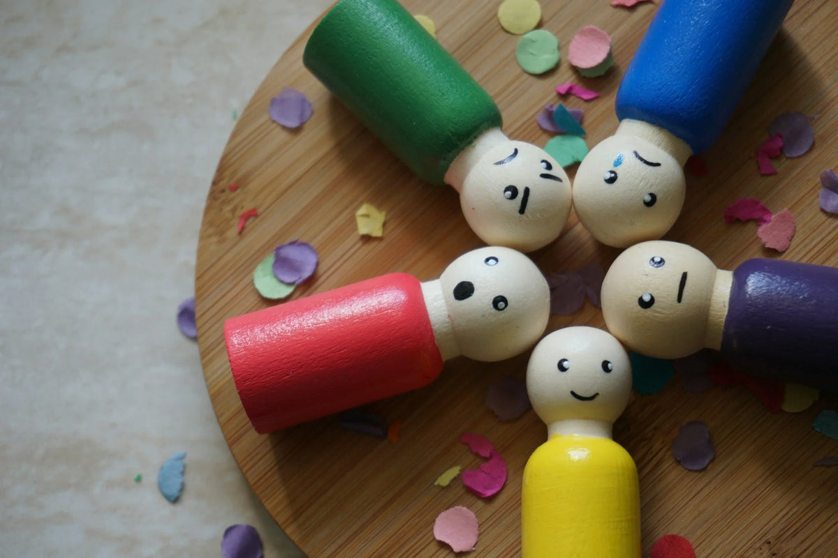 Five wooden peg dolls with different feelings painted on their faces, arranged in a circle on a wooden board